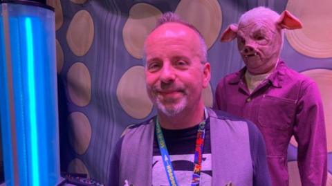 Sci-fi Scarborough's founder Steve Dickinson - a man with grey hair and a beard standing in front of a blue light and an extra-terrestrial model, which is wearing a pigs face mask covering their entire head and dark-coloured overalls.