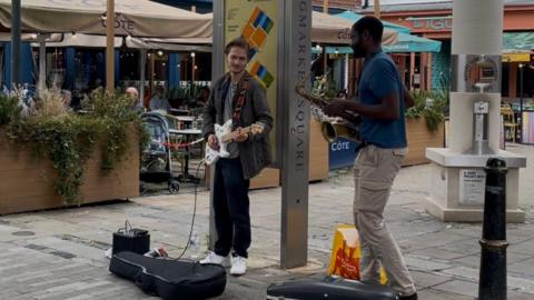 Two buskers, one playing an electric guitar and one playing a saxophone.