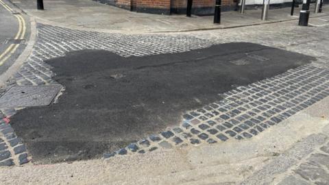 A section of a cobbled street which has had tarmac put on a section of it.