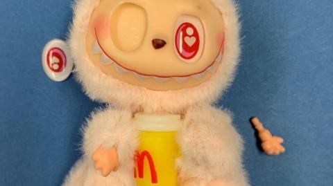Furry toy with eye missing and detached hand with a yellow and white beaker with a red letter M on it.