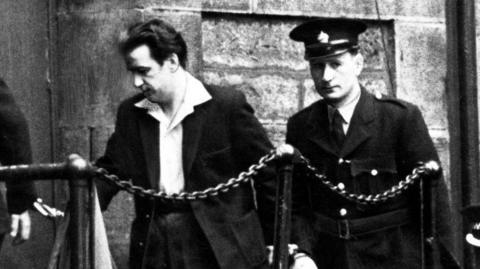 A black and white image from 1958 showing Peter Manuel - a man with thick dark hair in a dark suit with white, spread-collar, shirt - handcuffed to a prison officer, who is in uniform and looking at the camera. They are behind a chain fence and in front of a stone wall. The left arm of another officer can just be seen on the far left of frame. He too is handcuffed to Manuel.