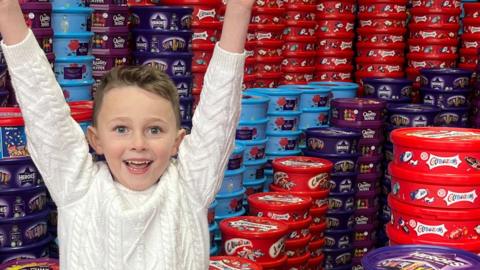 A young boy raises his arms in the air celebrating with a big smile on his face. He is wearing a white Aran sweater and is surrounded by chocolate tubs