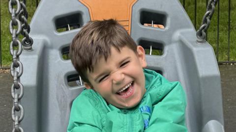 A boy in a green jack and brown hair sits on a grey and orange ergonomic chair swing at a playpark.