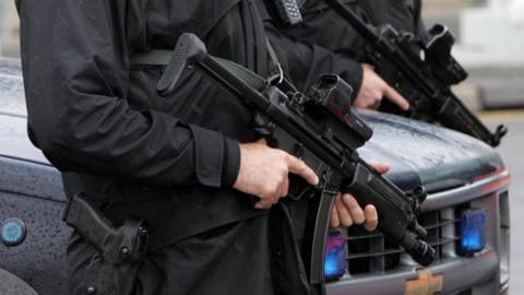 Two armed police officers stand in front of a black police car. The image is cropped close, showing them from the waist to the chest. Both are holding automatic-type weapons with the pistol of the one nearest the camera visible in a holster.