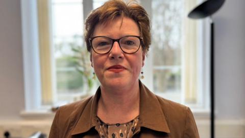 Jane Poole-Wilson has short brown hair and is wearing glasses. She has a brown patterned top under a brown jacket and is standing in an office in front of a window with a floor lamp to the right.