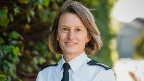A woman with shoulder length brown hair stands against a background of greenery and looks at the camera. She's wearing a police uniform white short-sleeved shirt with epaulettes and a dark tie with emblem.