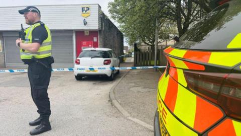 A police officer at a crime scene, with police tape and a police car also in shot.