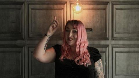 Debbie Todd has pink wavy hair. She is wearing red lipstick and a black top as she points to a light bulb shining above her head. Her right arm is covered in tattoos.