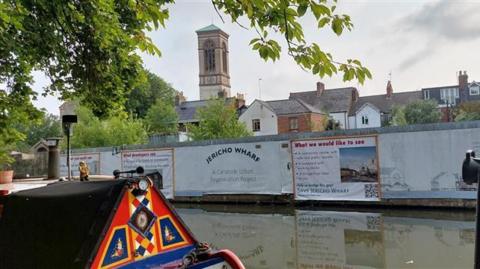 A view of the derelict site with tattered banners alongside the canal in Jericho. The church can be seen in the background. There is also a boat in the water.