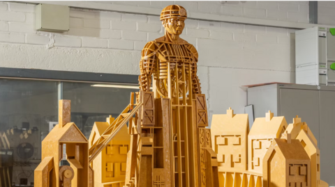 A model sculpture made of yellow wood depicts a miner rising out of some colliery buildings. He is wearing a miner's hat and lamp. 