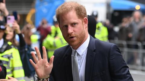 The Duke of Sussex looking waving while wearing a white-collared shirt, dark suit and grey tie