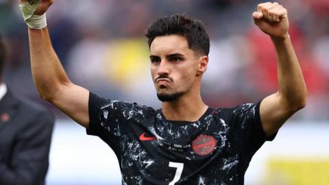 An image of Stephen holding both his fists up in the air defiantly after scoring the winning goal on Sunday. He is wearing a Black Canada Nike jersey which features a white pattern and a red Canada Soccer patch. He has short black hair and a black mustache and beard.