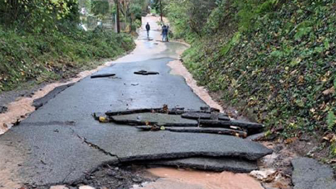 Road badly damaged by flooding, with the surface coming away, with flood water in the distance