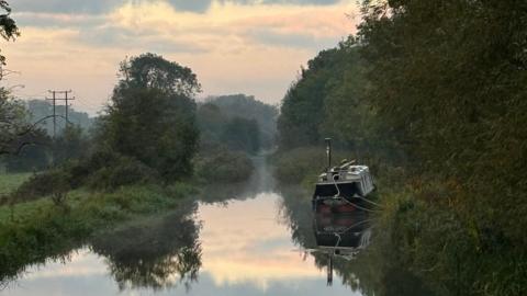 A narrowboat sits on a canal and is reflected in its smooth surface. Green trees and bushes line both banks. The sky has a pink tinge as the sun has just risen and there is an electricity pylon in the distance