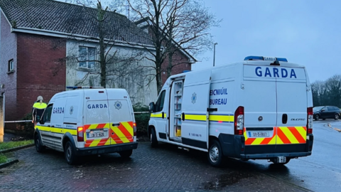 Two white Garda vans are parked side by side outside a residential property. The van on the left is smaller and a garda officer appears to be standing in front of the vehicle. The door on the larger van on the right has its sliding door open.