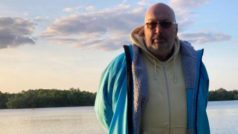 Martin Powers wearing a blue jacket with fluffy lining. He has stubble and is wearing glasses. He is standing in front of the large lake, which is lined with trees.