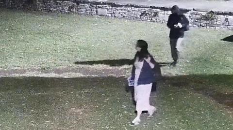 CCTV footage of a man and a woman walking through a park at night. There is a Cotswold stone wall at the edge of the park.
