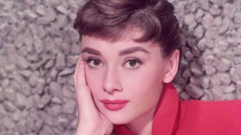 An image of a young Audrey Hepburn, with light brown hair and wearing a red jacket and red lipstick, with her face in her hand looking into the camera