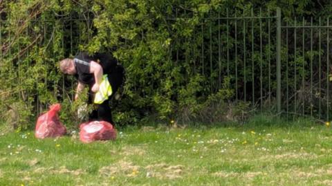 Paul Fairbridge collecting rubbish in a field. he is wearing a black shirt and a hi-vis jacket. He is bending down to collect rubbish. He has filled two red rubbish bags.