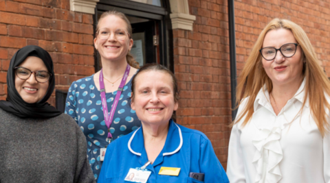 Four women are standing outside a brick building, containing a door, and looking at the camera. They are wearing glasses apart from the woman third from the left, who is in a blue nurse's uniform. The woman far left is in grey, the next person is wearing blue and the woman on the right is in white.