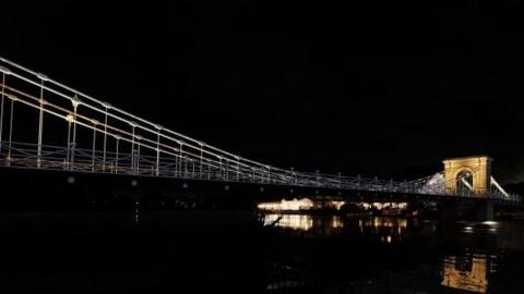 It is night time and a dimly lit suspension bridge crossing a river can be seen. The bridge viaduct can be seen illuminated at one end towards the background.