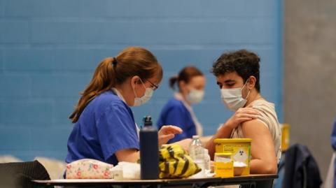 A nurse in scrubs and a mask prepares a vaccination at a table. A patient wearing a face mask has rolled up his sleeve.
