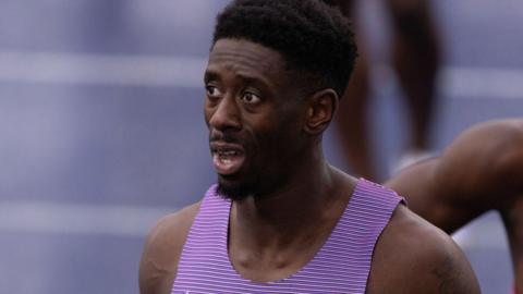 Close-up photograph of Reece Prescod following a 100m race. He has black hair and a goatee beard, and is wearing a purple running vest. Part of the track can be seen in the background.
