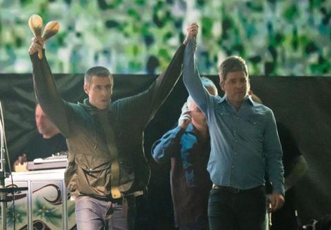 Photograph of Liam and Noel Gallagher standing hand in hand on stage.