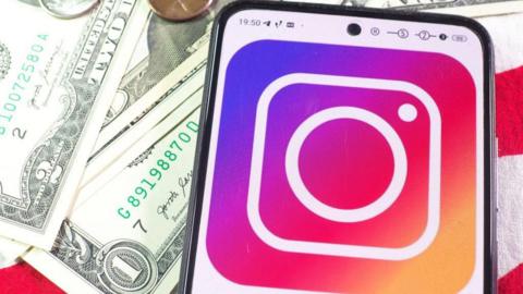 Instagram logo on a phone, which is lying on top of a stack of US dollar bills.