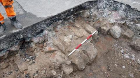 An image of a large hole dug into a concrete road, exposing rubble and bricks below the surface. A red and white striped stick marks out a section of the exposed bricks. To the left, stood on the concrete a pair of legs can be seen, wearing black work boots and florescent orange trousers. 