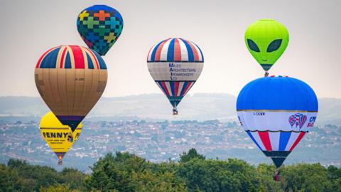 Six hot air balloons in the sky over Bristol. The city can be seen in the distance. The balloons all vary in shape and size. There is a green one shaped like an alien, two blue, red and white striped balloons, one with a union jack print on top, one multicoloured balloon and a yellow one. 