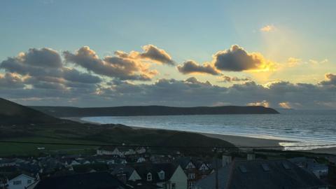 Small grey clouds float over a blue sky with the sun setting to the right and sea and houses in the foreground