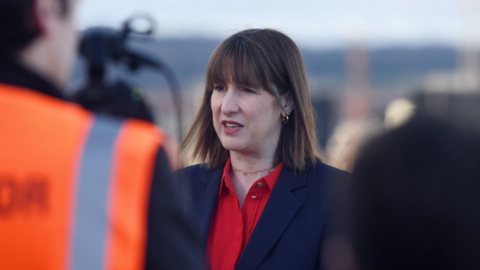 Chancellor Rachel Reeves speaks to reporters, wearing a scarlet open necked shirt, a navy suit jacket and a gold choker chain, with small gold hoop earrings. 