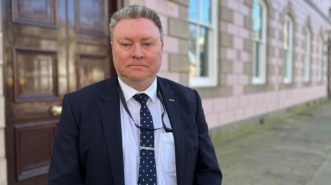 Jersey education minister Deputy Rob Ward stands outside a government building which has a large brown wooden door and lilac coloured brick work. He is wearing a dark suit and a white shirt with a black and white polka dot tie.
