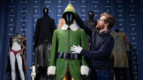 A man wear white gloves as he touches a black mannequin wearing a green coat with white fluffy cuffs and collar, yellow tights and a green and yellow pointy hat.