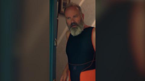 An older man with grey hair and beard wears a sleeveless, black and orange sporting catsuit. He is standing in a front doorway.