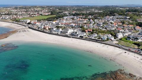 A drone picture showing Cobo bay in Guernsey. Blue water against white sandy beach with buildings and roads in the background. 