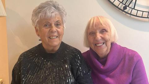 Two friends sit side by side and are smiling at the camera in this posed photograph. Barbara is wearing a black jumper embellished with sequins and Joyce is wearing a purple jumper. 
