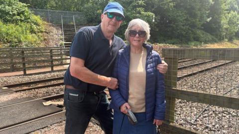A man in sunglasses, a baseball cap and dark polo shirt stands with his arm round a woman with blonde hair, a blue jacket, beige top and sunglasses. They are standing at the side of a railway line with pedestrian crossing mats across it.  
