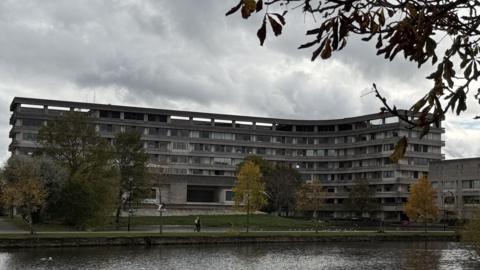 A large, curved concrete building with multiple rows of windows stands behind a grassy area and a pond. The building is Bedford Borough Council's headquarters. Several trees with autumn foliage are scattered across the scene, and branches frame the top of the image. The sky is overcast, creating a moody atmosphere, and a few people can be seen walking along the path near the water.