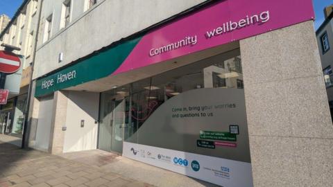 A general view of the Hope Haven hub. The premises are on the ground floor at the corner of a street. The front has large shop-style windows, partially frosted. A sign reads Hope Haven on a green and pink background.