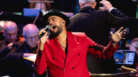 A man singing at the BBC Proms with an orchestra and conductor behind him.