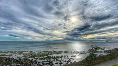 Blue, yellow and grey clouds fill the sky as the sun rises over silvery sea