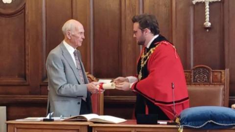 Peter Isaac, who has short white hair and is wearing a grey suit, accepts a roll of paper that has been bound with red ribbon from the Mayor of Hereford, Dan Powell, who is wearing red robes and a gold chain. Behind them are wooden panelled walls and a leather chair.