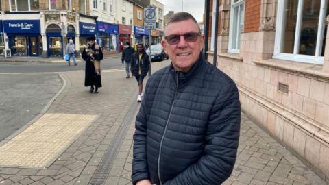 Tom Ryder is wearing glasses and a navy coat. He is smiling at the camera and has short hair.  There are shops in the backgroud