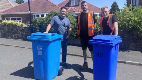 Three men are stood in a street with two large blue wheelie bins. The man on the left has dark curly hair, glasses and is wearing a blue t shirt and jeans. The other two men are wearing black t-shirts, high vis orange vests and shorts