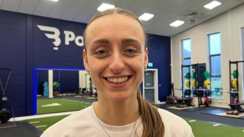 The photo shows a woman smiling at the camera. She has brown hair which has been tied back into a ponytail. She's wearing a white top and a silver necklace. She's standing in a gym-like room. There are weights to her right, and a mirror to her left. 