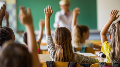 A group of young school children sat in a class with their hands in the air, viewed from behind, with a teacher out of focus. 
