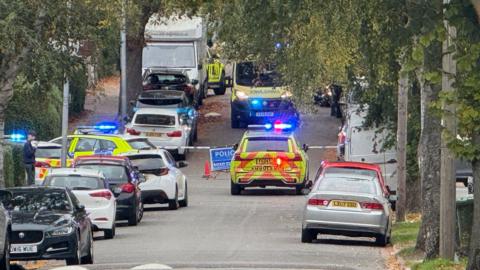An uphill road lined with parked cars. In the distance are multiple emergency vehicles with flashing lights. A blue cordon and a blue sign reading "POLICE ROAD CLOSED" sits in the middle of the road.
