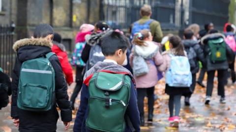 The image shows a group of schoolchildren walking along a pavement covered with leaves, wearing backpacks and coats. They are heading in the same direction towards a school entrance, with an adult walking ahead of them. Tall black railings and brick buildings line the street.